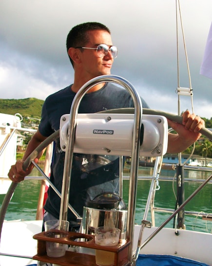 A person wearing sunglasses is steering a boat, with lush green hills and cloudy skies in the background. The boat features a steering wheel and navigation equipment branded 'NavPod'. There are glasses with drinks on a wooden holder attached to the equipment.