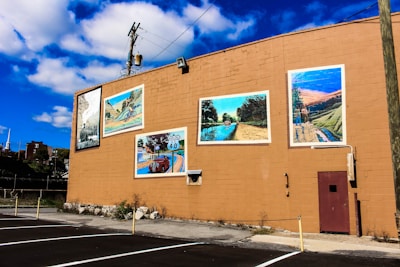 A large beige wall with several colorful murals depicting different scenes, including a landscape, a body of water, an old car, and a scenic road. The wall is part of a larger building with a small red door, and the area in front of it is a paved parking lot with a few yellow poles. The sky above is blue with scattered white clouds.