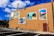 A large beige wall with several colorful murals depicting different scenes, including a landscape, a body of water, an old car, and a scenic road. The wall is part of a larger building with a small red door, and the area in front of it is a paved parking lot with a few yellow poles. The sky above is blue with scattered white clouds.