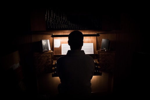 A person is seated at an organ, back turned to the viewer, in a dimly lit room. Sheet music and books are visible on the organ stand. The overall atmosphere is quiet and intimate, with a strong focus on the light illuminating the music.