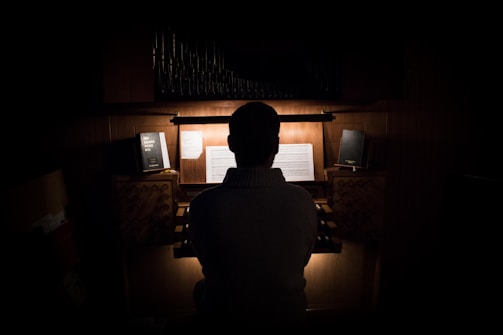 A person is seated at an organ, back turned to the viewer, in a dimly lit room. Sheet music and books are visible on the organ stand. The overall atmosphere is quiet and intimate, with a strong focus on the light illuminating the music.