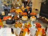 An assortment of colorful pastries and cakes is displayed behind a glass window in a bakery. The display includes various cakes topped with fresh fruits like strawberries and citrus slices. There are also chocolate cakes and tarts arranged neatly. In the background, a person is seen preparing more baked goods.