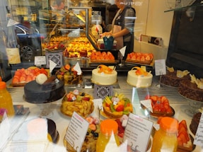 An assortment of colorful pastries and cakes is displayed behind a glass window in a bakery. The display includes various cakes topped with fresh fruits like strawberries and citrus slices. There are also chocolate cakes and tarts arranged neatly. In the background, a person is seen preparing more baked goods.