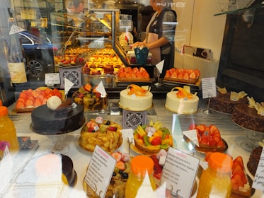 An assortment of colorful pastries and cakes is displayed behind a glass window in a bakery. The display includes various cakes topped with fresh fruits like strawberries and citrus slices. There are also chocolate cakes and tarts arranged neatly. In the background, a person is seen preparing more baked goods.