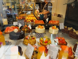 An assortment of colorful pastries and cakes is displayed behind a glass window in a bakery. The display includes various cakes topped with fresh fruits like strawberries and citrus slices. There are also chocolate cakes and tarts arranged neatly. In the background, a person is seen preparing more baked goods.
