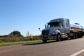A large tanker truck drives along a rural road, surrounded by grassy fields and distant trees. The sky is clear and blue.