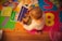 A small child is seated on a colorful foam play mat engaged with a book titled 'Moonbeam Bear.' Several other children's books are scattered around. The flooring underneath the mat is wooden, and there is a pink basket nearby.