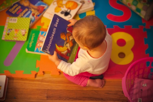 A small child is seated on a colorful foam play mat engaged with a book titled 'Moonbeam Bear.' Several other children's books are scattered around. The flooring underneath the mat is wooden, and there is a pink basket nearby.