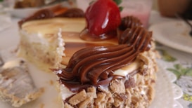 A close-up view of a slice of cake topped with a glossy red strawberry and swirled chocolate icing. The cake appears to have a creamy layer with a nutty, crumbly crust. In the background, a blurred setting suggests a dining arrangement.
