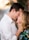 A couple stands closely, touching foreheads and smiling warmly at each other. The man wears a white shirt, while the woman has blonde hair and wears a colorful floral outfit. Their hands gently touch each other's faces, conveying intimacy and affection. The background is softly blurred, focusing attention on the couple.