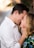 A couple stands closely, touching foreheads and smiling warmly at each other. The man wears a white shirt, while the woman has blonde hair and wears a colorful floral outfit. Their hands gently touch each other's faces, conveying intimacy and affection. The background is softly blurred, focusing attention on the couple.