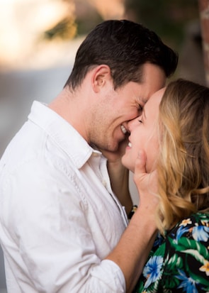 A couple stands closely, touching foreheads and smiling warmly at each other. The man wears a white shirt, while the woman has blonde hair and wears a colorful floral outfit. Their hands gently touch each other's faces, conveying intimacy and affection. The background is softly blurred, focusing attention on the couple.