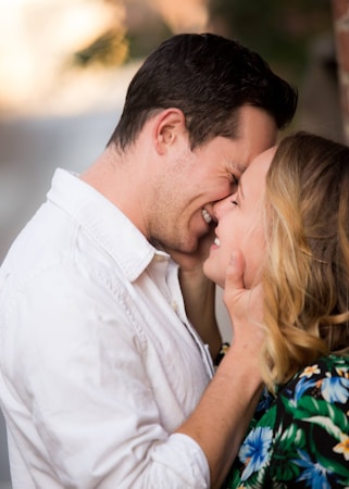 A couple stands closely, touching foreheads and smiling warmly at each other. The man wears a white shirt, while the woman has blonde hair and wears a colorful floral outfit. Their hands gently touch each other's faces, conveying intimacy and affection. The background is softly blurred, focusing attention on the couple.
