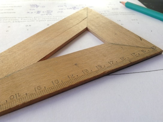 A wooden triangular ruler with metric measurements is placed on top of some paper with mathematics calculations. A teal pencil is visible in the background.