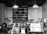 A coffee shop counter with a woman behind it, surrounded by various equipment like coffee grinders, an espresso machine, and cups. The menu board behind displays coffee and tea options with prices. Overhead lights hang down, and various jars and containers are arranged on the counter.