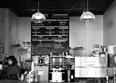 A coffee shop counter with a woman behind it, surrounded by various equipment like coffee grinders, an espresso machine, and cups. The menu board behind displays coffee and tea options with prices. Overhead lights hang down, and various jars and containers are arranged on the counter.