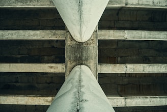 A close-up view of the underside of a concrete bridge, showcasing the large beams and the weathered texture of the structure. The perspective emphasizes the symmetry and the intersecting lines of the beams, with shadows adding depth to the image.