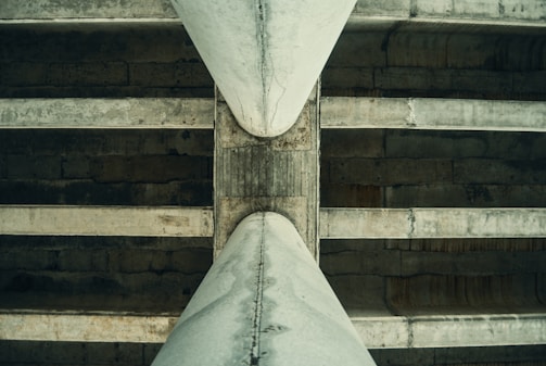 A close-up view of the underside of a concrete bridge, showcasing the large beams and the weathered texture of the structure. The perspective emphasizes the symmetry and the intersecting lines of the beams, with shadows adding depth to the image.