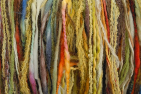 A close-up of various strands of yarn in multiple colors, including shades of yellow, green, red, blue, and brown. The yarn is textured, with some strands appearing thick and others thin, creating a layered and vibrant appearance.