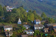 A small rural village nestled among lush green hills and dense forests. Several houses with slate roofs are scattered along the hillside, surrounded by trees and vegetation. A white temple with a pointed roof stands prominently in the center of the village, adding a cultural touch. The crisp morning light casts shadows that emphasize the contours of the landscape.