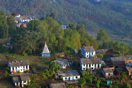 A small rural village nestled among lush green hills and dense forests. Several houses with slate roofs are scattered along the hillside, surrounded by trees and vegetation. A white temple with a pointed roof stands prominently in the center of the village, adding a cultural touch. The crisp morning light casts shadows that emphasize the contours of the landscape.