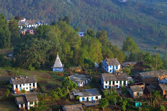 A small rural village nestled among lush green hills and dense forests. Several houses with slate roofs are scattered along the hillside, surrounded by trees and vegetation. A white temple with a pointed roof stands prominently in the center of the village, adding a cultural touch. The crisp morning light casts shadows that emphasize the contours of the landscape.
