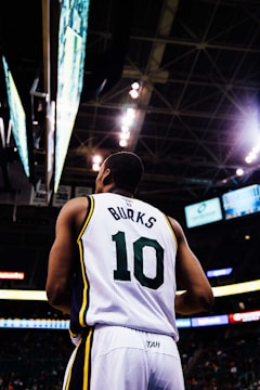 A basketball player wearing a white jersey with the number 10 and the name 'Burks' stands on a basketball court. The arena's ceiling and lights are visible above, along with electronic screens displaying game information.
