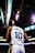 A basketball player wearing a white jersey with the number 10 and the name 'Burks' stands on a basketball court. The arena's ceiling and lights are visible above, along with electronic screens displaying game information.