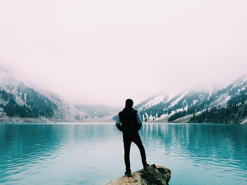 man standing on brown rock facing beside body of water