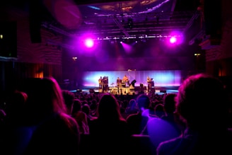 A dimly lit concert venue features a band performing on stage under vibrant purple and pink stage lights. The audience is seated in front of the stage, silhouetted against the bright lights. The stage is equipped with musical instruments, including drums and guitars, and the backdrop glows with soft, colorful lighting that enhances the atmosphere.