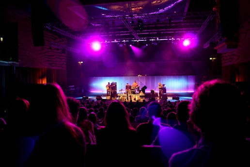 A dimly lit concert venue features a band performing on stage under vibrant purple and pink stage lights. The audience is seated in front of the stage, silhouetted against the bright lights. The stage is equipped with musical instruments, including drums and guitars, and the backdrop glows with soft, colorful lighting that enhances the atmosphere.