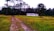 A rural landscape featuring a dirt path leading to a small, white, rectangular building with a metal roof, set amidst grassy fields. Tall trees form a dense forest backdrop under an overcast sky.