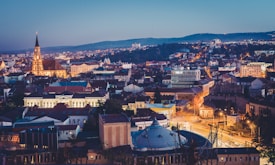 An aerial view of a cityscape during dusk with a prominent church featuring a tall spire illuminated in the center. Surrounding buildings with a mix of traditional and modern architecture are lit with warm streetlights. The background includes rolling hills under a darkening sky.