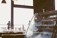 The interior of a cafe or restaurant with a focus on a display case filled with various desserts. The room is softly lit with hanging lamps and there are a few people visible outside through large windows, suggesting an airy and open atmosphere. On the table, there are condiments and utensils, adding to the casual dining feel.