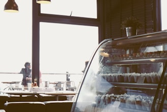 The interior of a cafe or restaurant with a focus on a display case filled with various desserts. The room is softly lit with hanging lamps and there are a few people visible outside through large windows, suggesting an airy and open atmosphere. On the table, there are condiments and utensils, adding to the casual dining feel.