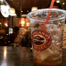 A close-up of a plastic cup with a Highland Coffee logo, partially filled with a cold beverage and ice, on a table in a dimly lit cafe. A red straw is inserted into the cup. In the background, warm lighting and interior details of the cafe are visible, with other patrons sitting and a blurred cafe display board.