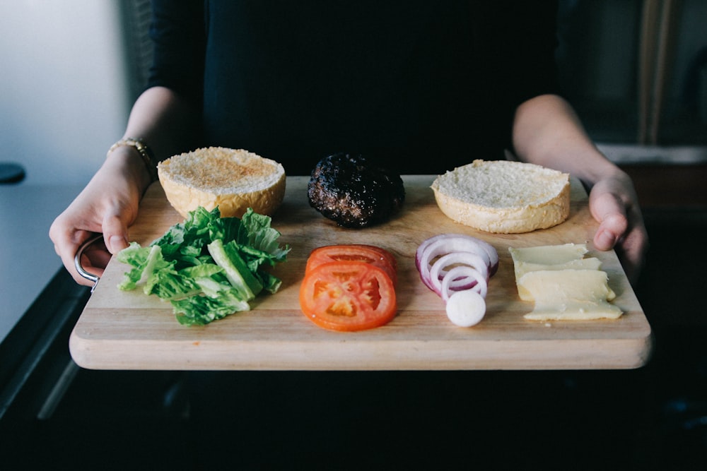 woman holding up wooden tray with lettuce, tomato, onions rings and cooked patty for burger