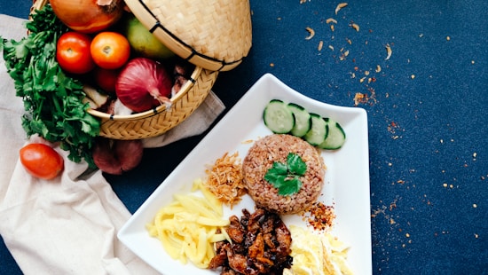 A vibrant dish presented on a white plate features brown rice garnished with a sprig of cilantro, slices of cucumber, and assorted accompaniments such as shredded vegetables and diced cooked meat. A wicker basket beside the plate is filled with fresh produce including tomatoes, onions, and greens, all arranged on a textured blue surface scattered with spice flakes.