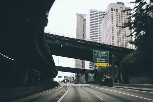 A road leading into a city, flanked by tall buildings. Overhead, there are multi-level overpasses. In the foreground, a road sign provides directions to Bellevue and Spokane via Interstate 90. The scene conveys a typical urban environment with towering skyscrapers and infrastructure.