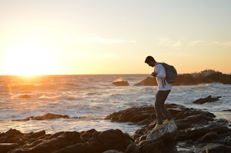 A joyful traveler standing on a rocky shore, watching playful otters swim near the waves under a golden sunset.