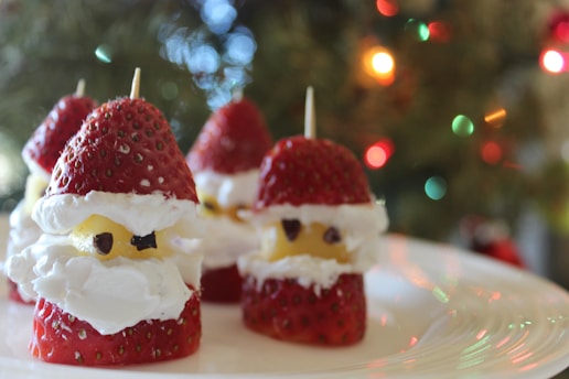 Several strawberries are decorated with cream and small chocolate pieces to resemble Christmas-themed characters, placed on a plate with blurry, colorful lights in the background.