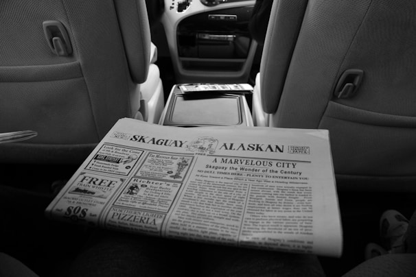 A black and white newspaper titled 'Skaguay Alaskan' is being held in the interior of a car. The newspaper features various headlines and advertisements. The car interior includes visible front seats and a center console.