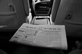 A black and white newspaper titled 'Skaguay Alaskan' is being held in the interior of a car. The newspaper features various headlines and advertisements. The car interior includes visible front seats and a center console.