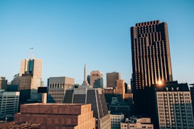 A city skyline at sunset with various modern skyscrapers and office buildings. The sun reflects off the windows of a tall black building on the right, creating a warm glow. The sky is clear with a pale blue color, enhancing the golden hues of the sun.