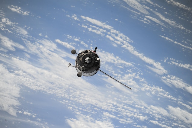 A spacecraft is orbiting above the Earth, surrounded by a backdrop of the planet's atmosphere and scattered clouds. The spacecraft appears to be metallic with various protruding antennas and equipment, reflecting sunlight.