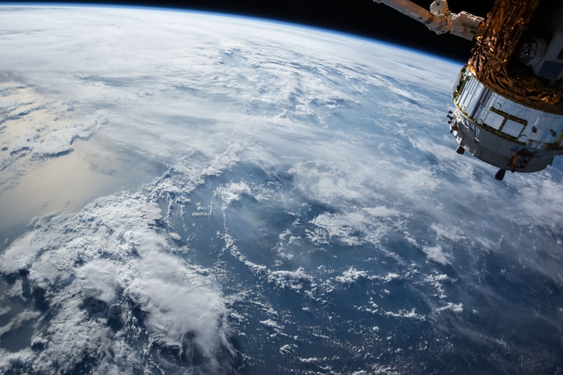 A view of Earth from space showing cloudy skies over a vast expanse of ocean, with a spacecraft partially visible on the right side of the image.