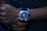 A hand with a stylish wristwatch featuring a dark, sleek design and silver accents. The wrist is adorned with a bracelet composed of white and black beads, creating a contrast with the watch. The person appears to be wearing a dark textured shirt in the background, casting an overall dim and sophisticated ambiance.