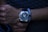 A hand with a stylish wristwatch featuring a dark, sleek design and silver accents. The wrist is adorned with a bracelet composed of white and black beads, creating a contrast with the watch. The person appears to be wearing a dark textured shirt in the background, casting an overall dim and sophisticated ambiance.