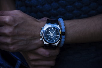 A hand with a stylish wristwatch featuring a dark, sleek design and silver accents. The wrist is adorned with a bracelet composed of white and black beads, creating a contrast with the watch. The person appears to be wearing a dark textured shirt in the background, casting an overall dim and sophisticated ambiance.
