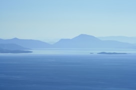 A serene landscape featuring a calm body of water with gentle waves. In the distance, layers of mountains rise, creating a sense of depth. The scene is enveloped in soft, muted blue tones, suggesting a peaceful, tranquil atmosphere.